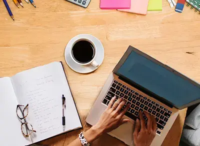 Laptop on a desk with hands typing, next to a notebook, pen, glasses, coffee cup, and colorful sticky notes.