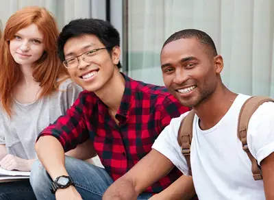 Three students seated together outdoors, smiling and wearing casual clothing with backpacks in front of a campus building.