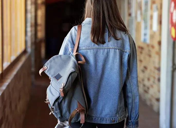 Person wearing a denim jacket with a blue backpack, seen from behind in a softly lit hallway.