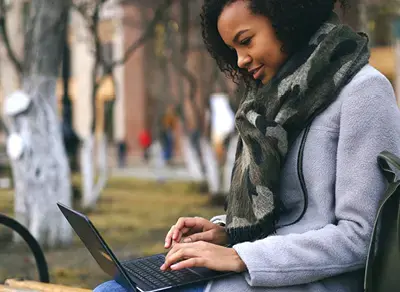 Person seated outdoors typing on a laptop, wearing a coat and scarf, with a blurred campus setting in the background.