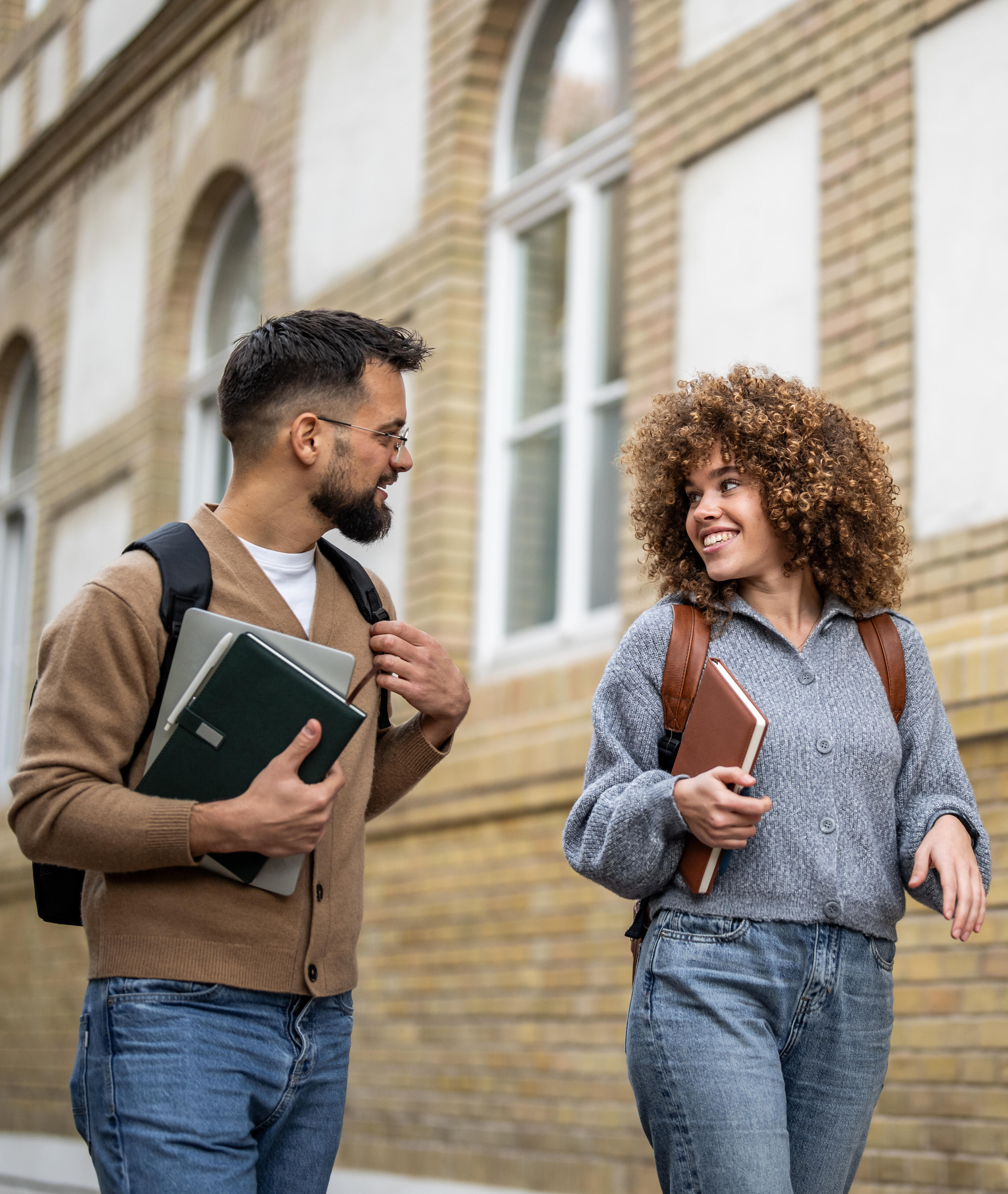 Two young adult students are walking and talking outside of a brick university building, carrying books and backpacks, enjoying their time on campus
